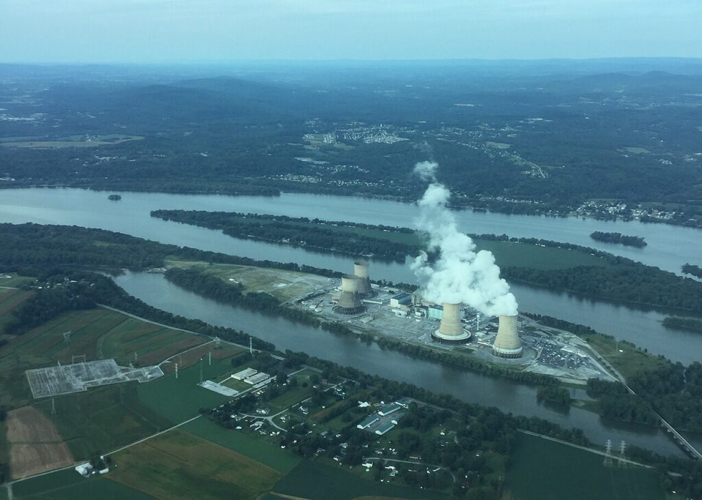 Three Mile Island, a nuclear power plant, and nearby Goldsboro in Pennsylvania. The plant sits on an island surrounded by waterways and greenery. The photo is taken from the air. Plumes of water vapour rise from the cooling towers