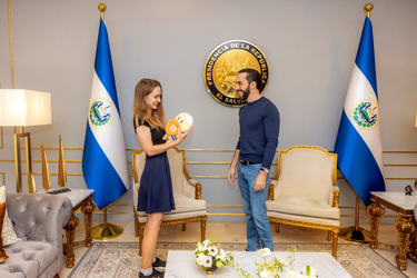 Photograph of two people standing facing each other in a room which has armchairs, two El Salvadorean flags on upright poles, and also an El Salvadorean presidential seal on the wall.
At left is a smiling woman with long hair, wearing a dress. She is holding a soft toy figure which is in turn holding a soft orange disc with a Bitcoin logo on it.
At right is a smiling bearded man wearing a sweater and jeans.