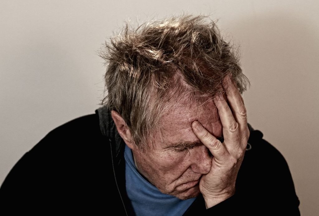 Head and shoulders photograph of a man who looks exhausted, holding his head in his left hand, with his eyes closed.