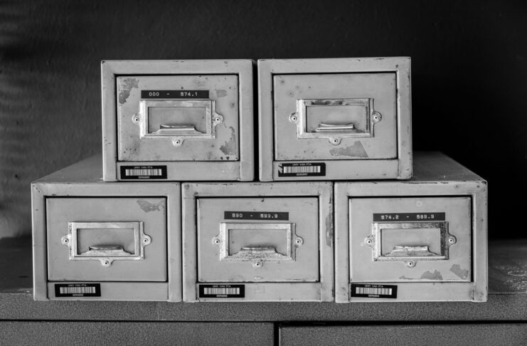 Black and white photograph of five old metal file boxes such as might contain card indexes. The boxes are stacked in two rows, with a lower row of three boxes and a row of two on top.
