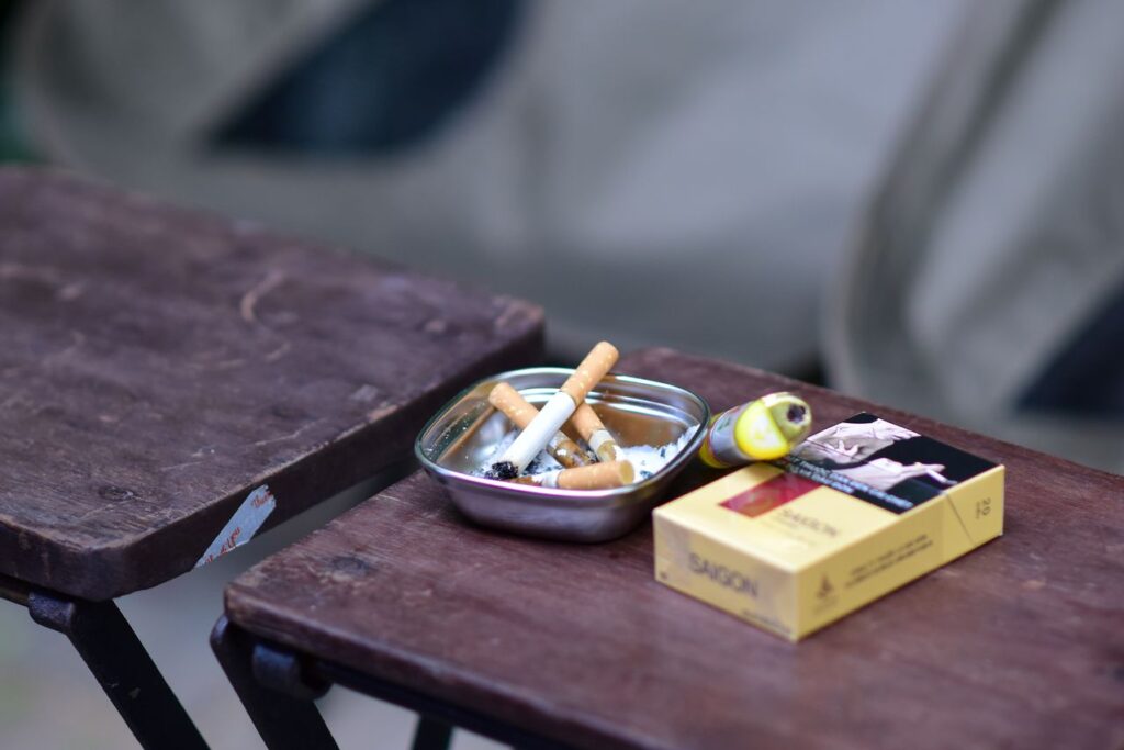 Photograph of an ashtray containing several cigarette butts, next to a pack of cigarettes and a lighter on a small wooden table.