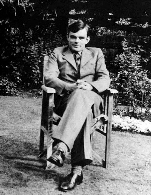 Black and white photograph of a young man sat on a wooden armchair in a garden. He is clean shaven, and wearing a suit and tie. His legs are crossed and his hands are folded together on his knee.