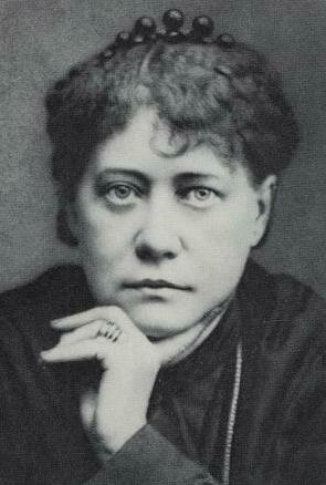 A black and white photo of a serious-looking woman looking face-on at the camera, resting her chin on her hand. She has dark, curly hair that's put up with a headband and wears a series of rings on her middle finger