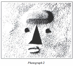 Black and white photograph captioned "Photograph 2". Against a bright white background speckled with grey are features resembling a face. Two deep circles of black form the eyes, a crisp triangular shadow forms the nose, and an oval ring of shadow forms a mouth.