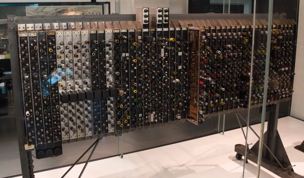 Photograph of a rack of electronic equipment, mounted on a wheeled stand.
Approximately 45 vertical channels each contain ten or twelve pairs of cylindrical plugs or end caps of components within the channel.
One of the channels has been pulled out, showing electrical circuitry within.