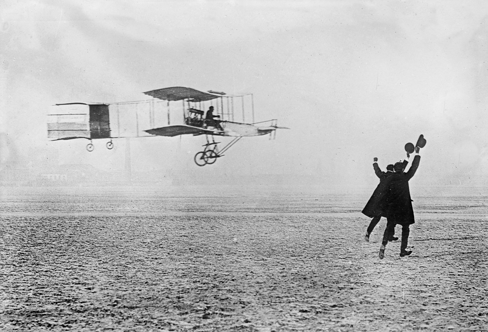Grainy black and white photograph of a rudimentary biplane flying above clear level ground at an altitude of a few metres. In the foreground, two spectators on the ground are waving their hats in the air, as if in celebration.