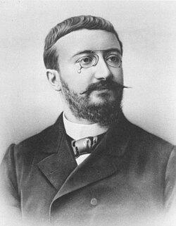 Black and white portrait photograph of a man with short hair, a short beard and moustache. He is wearing a cravat, and shirt with a stiff collar under a double-breasted jacket. His moustache tapers to narrow probably waxed points and he is wearing pince nez glasses.