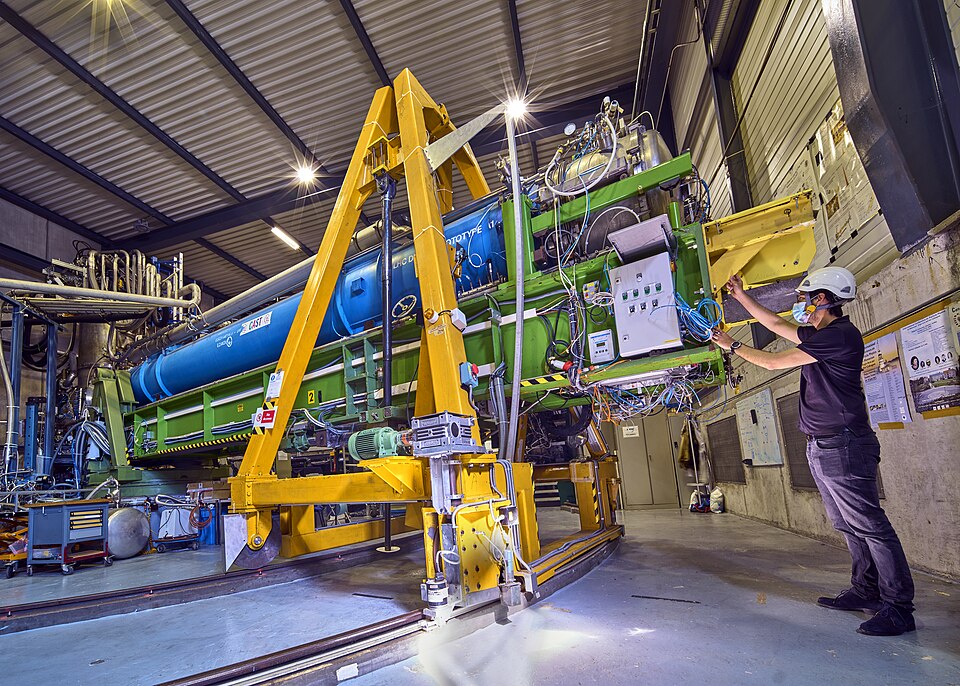 Photograph showing a large assembly of equipment comprising cables and pipes which must weigh several tonnes, on a wheeled gantry which can move on rails. A man wearing a surgical mask and hard hat is working at one end of the machine near the camera. The whole assembly is approximately twice the height of the man and extends perhaps 15 metres away from him, where it connects to another set of large pipes.