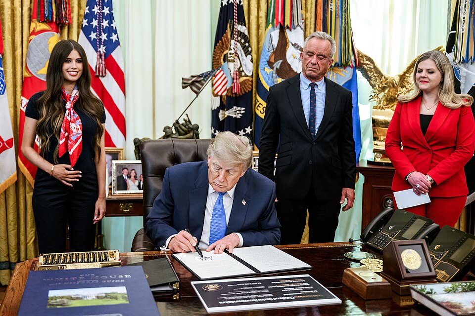 Photograph taken in Oval Office. Donald Trump signs papers on his desk, as RFK junior stands watching.