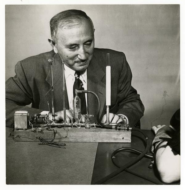 Black and white photograph of a middle-aged man wearing suit and tie. He is sat at a table, on which is an apparatus comprising several metal and glass tubes, some wires and a small box which could be battery. The apparatus is connected by a tube to a cuff secured on the wrist of someone sat just out of shot, whose forearm we can see resting on the table.