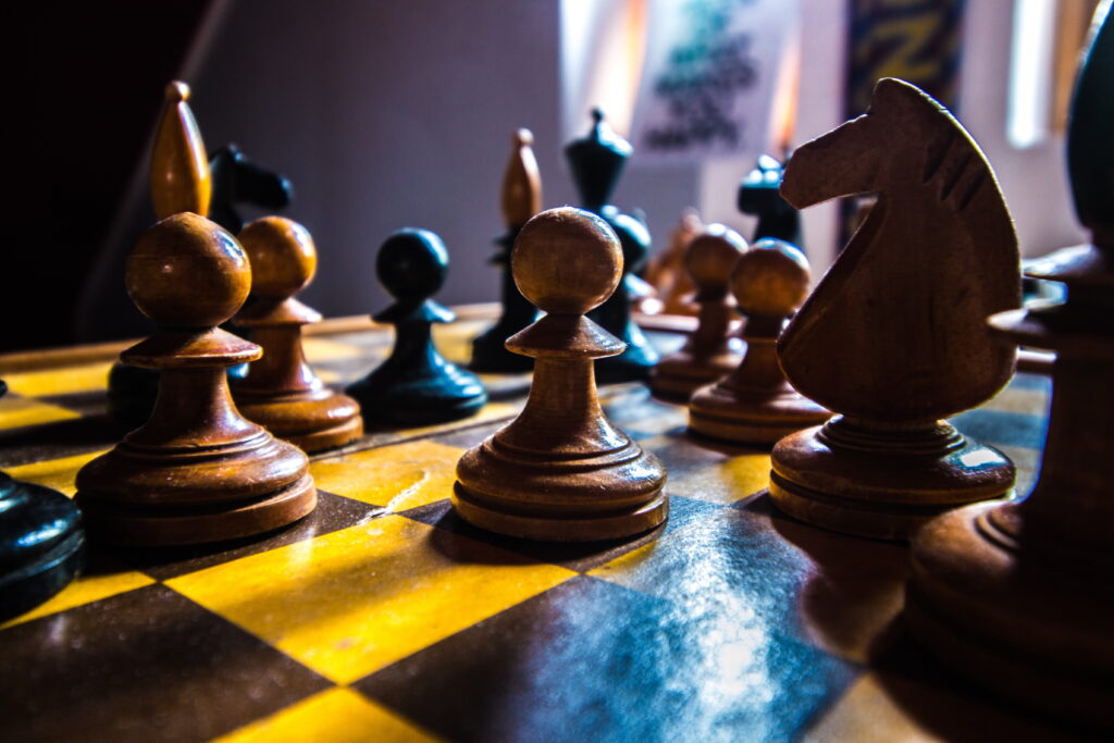 Close-up photo of a chess board with pieces standing on it, taken from near the board's surface. In the background there's a window where natural light is coming from to illuminate the scene and cast the pieces nearest the camera in relative darkness.