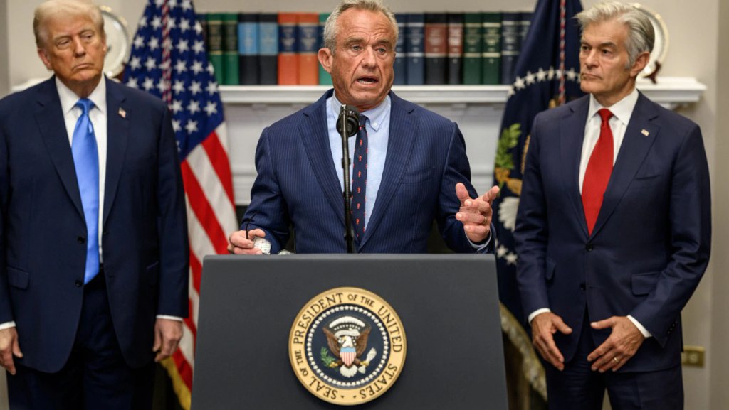 RFK junior speaking at a lectern bearing the US Presidential seal. Standing beside him and looking on are Donald Trump and Mehmet Oz.