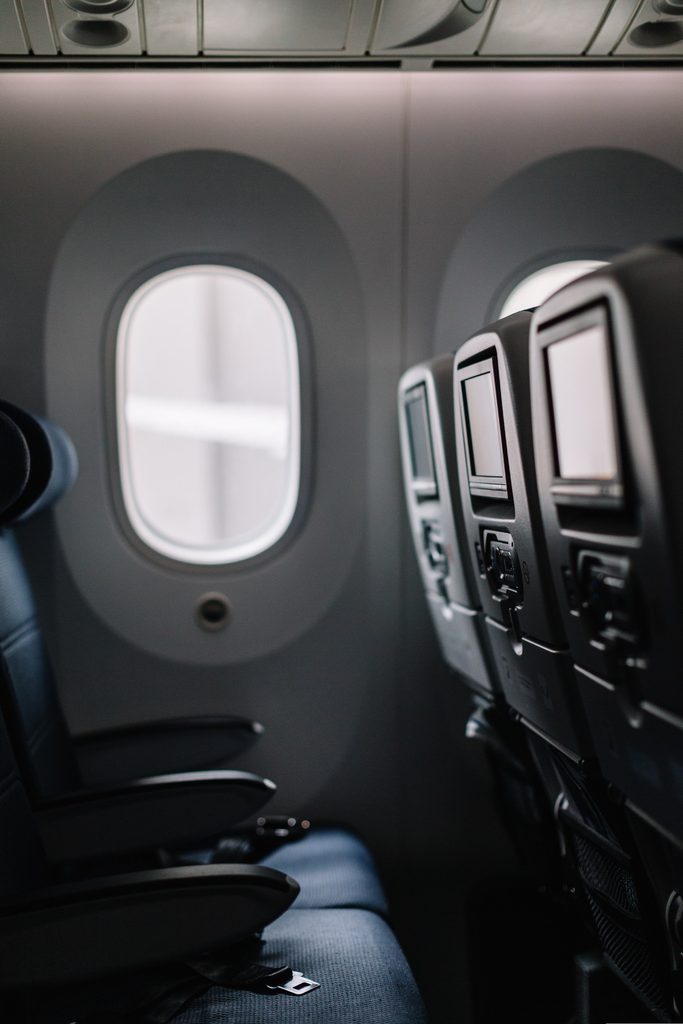 Black-and-white photograph of empty high-end plane seats, looking from a right-hand row across three seats to the left at the opposite window. The seats have screens in the backs of the headrests.